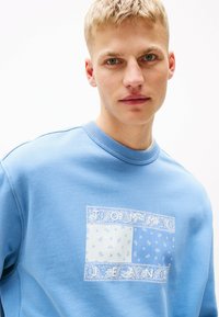 Sudadera de color azul claro con un gráfico central que presenta patrones de paisley blancos y el texto "TOMMY JEANS" en un diseño rectangular. Textura suave.