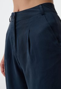 Marineblaue, maßgeschneiderte Hose mit strukturiertem Finish, versehen mit Eingrifftaschen und einem Knopfverschluss. Der flache Bund betont das Design.