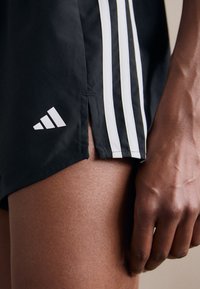 Black athletic shorts featuring white side stripes, a slit at the hem, and an Adidas logo on the lower left side. Smooth texture.