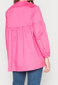 Blusa rosa a maniche lunghe con corpetto arricciato, scollatura rotonda e polsini con bottoni, abbinata a jeans blu. Tessuto morbido e leggero.