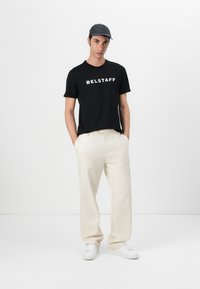 Black t-shirt with white "BELSTAFF" logo, cream-colored pants, and white sneakers. The model wears a dark cap, standing against a plain background.
