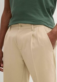 Pantaloni beige a pieghe con frontale liscio, chiusura con bottone e tasche laterali. Il materiale appare liscio con una vestibilità sartoriale.
