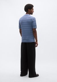 Μπλε πλεκτό πόλο μπλουζάκι με υφασμένο γεωμετρικό σχέδιο, κοντά μανίκια, σετ με μαύρο παντελόνι φαρδιάς γραμμής. Πλάγια όψη από πίσω.