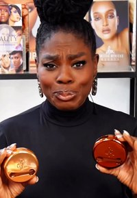 To runde beholdere holdt, en gull og en bronse. Begge har glatte overflater med et preget logo som reflekterer lys. Svart bakgrunn.