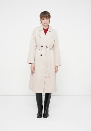 Gräddfärgad trenchcoat med bälte, dubbelknäppt design, stora svarta knappar och breda ärmar, i kombination med åtsittande svarta stövlar.