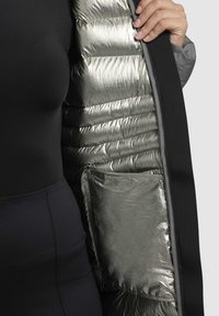 Giubbotto imbottito nero con fodera riflettente argento, caratterizzato da una texture trapuntata e una tasca laterale. Il materiale esterno sembra liscio.