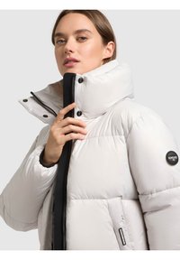 Giacca a piumino bianca leggera con collo alto, cerniera nera e bottoni a pressione. Presenta una toppa con marchio sulla manica e una texture trapuntata.