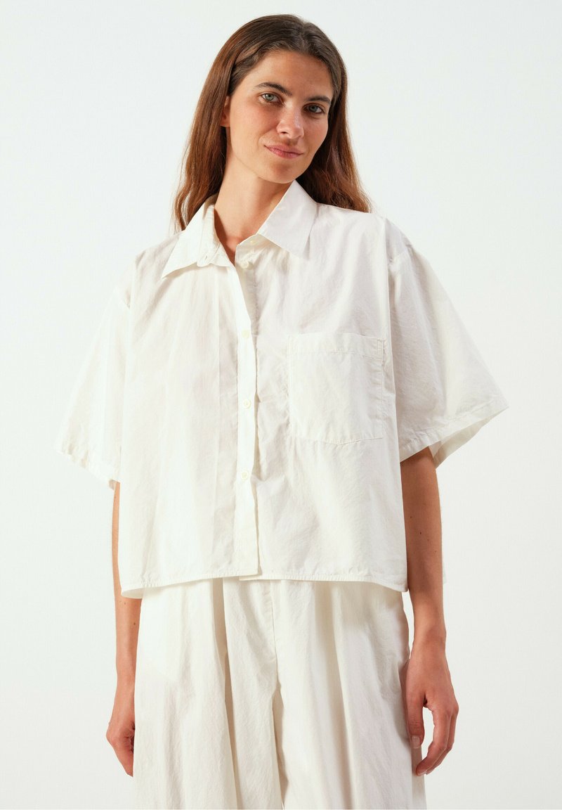 Jeune femme aux longs cheveux bruns portant une chemise blanche ample à manches courtes boutonnée et un pantalon large assorti sur un fond uni.