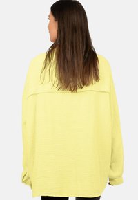 Blusa gialla oversize in tessuto texturizzato e stropicciato. Presenta un giromanica posteriore e maniche scese, con maniche lunghe e orlo arrotondato.