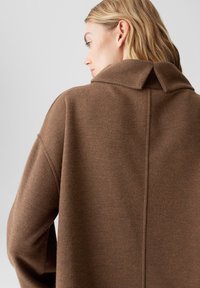 Cappotto in lana marrone con colletto strutturato, vestibilità comoda e dettagli di cucitura visibili lungo la cucitura posteriore. Texture morbida, design minimale.