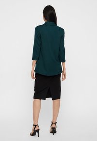 Blazer teal a tre quarti, gonna nera al ginocchio con spacco laterale e sandali neri con cinturini. Tessuto liscio con vestibilità su misura.