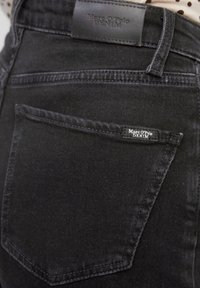 Jeans de mezclilla negra con un tejido texturizado, un bolsillo trasero con una pequeña etiqueta de marca y un parche de cuero en la cintura.