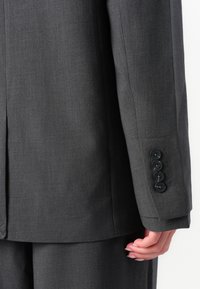 Ärmel und Knöpfe einer dunkelgrauen Anzugjacke auf dem Arm einer Person, die Hand entspannt neben dem Körper, um die Textur des Stoffes und die Nähte zu zeigen.