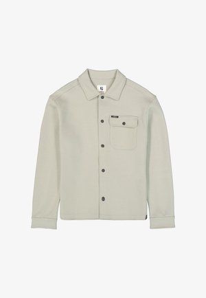 Veste-chemise beige clair avec manches longues, poche poitrine à bouton unique et petite étiquette de marque noire sur la poche.