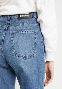 Blå denimjeans med en bakficka, synlig söm och en svart etikett med texten "DRDENIM" vid midjan. Tyget har en strukturerad yta.