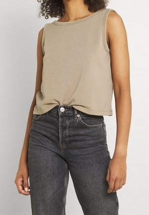 Vrouw die een mouwloos beige topje draagt, lichtjes ingestopt in een hooggetailleerde vervaagde zwarte spijkerbroek, staand tegen een effen achtergrond.