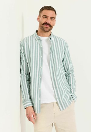 Man with mustache leans against wall wearing green and white striped shirt over white t-shirt, beige pants, tattoos visible on left hand.