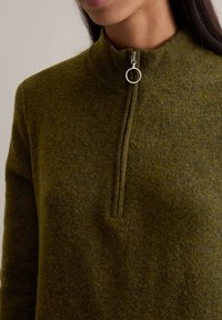 Olivgrüner Wollpullover mit hohem Kragen und einem Reißverschluss vorne, ausgestattet mit einem silbernen Ring-Zipper und einem strukturierten, weichen Stoff.