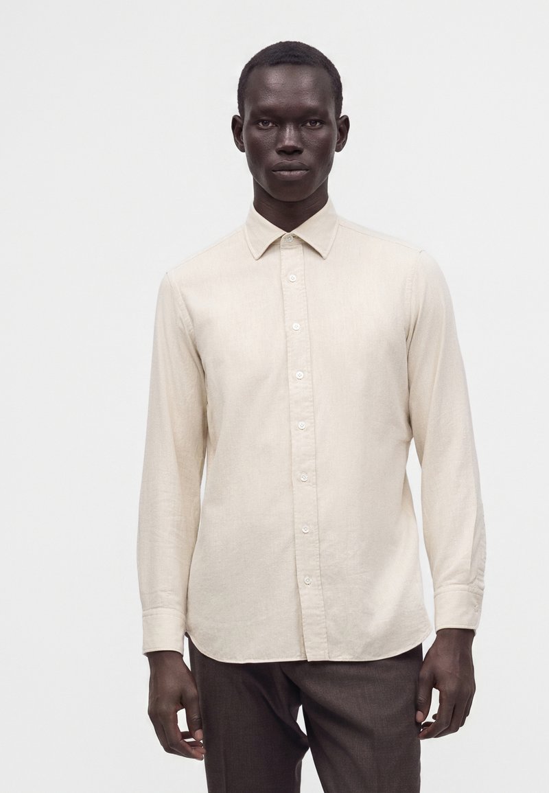 Jeune homme à la peau foncée portant une chemise beige à manches longues boutonnée et un pantalon foncé, debout devant un fond blanc uni.