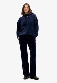 Vrouw staat in een marineblauwe velours hoodie met ritssluiting en bijpassende wijde broek met handen in de zakken tegen een witte achtergrond.