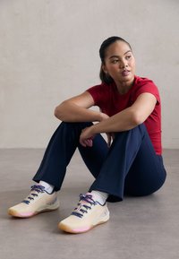 Męskie sneakersy sportowe w jasnym beżu z różowymi akcentami, siateczkową cholewką i granatowymi sznurówkami. Model nosi czerwony T-shirt i granatowe spodnie.
