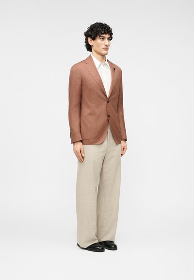 Καφέ λινό σακάκι με δύο μπροστινές τσέπες, συνδυασμένο με ανοιχτό μπεζ παντελόνι φαρδιάς γραμμής και μαύρα παπούτσια. Λευκό πουκάμισο με γιακά από μέσα.