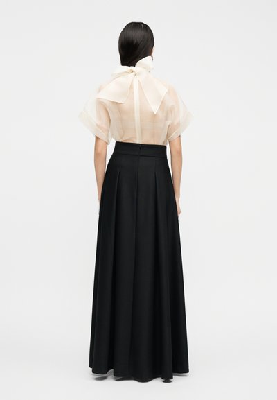 Femme aux longs cheveux noirs portant un chemisier blanc transparent avec un grand nœud au niveau du cou et une jupe noire longue taille haute, vue de dos.