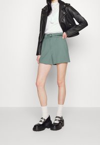 Pantaloni shorts verde khaki con vita cinturata, abbinati a una blusa bianca con dettagli in pizzo e un giubbotto di pelle nera. Scarpe con plateau nere.
