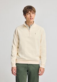 Sudadera de forro polar beige con cuello de cremallera de cuarto, puños acanalados y un pequeño logo en el pecho. Lleva puesta sobre una camisa de rayas con cuello.