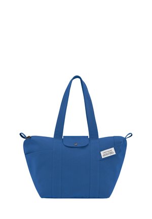 Blue canvas tote bag with two long handles, a button closure, and a side pocket featuring a label, structured and spacious design.