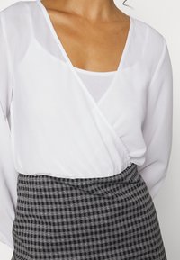 Blusa branca envolvente com mangas longas, combinada com uma saia ajustada de padrão xadrez preto e branco. A blusa tem um decote em V profundo e uma textura lisa.