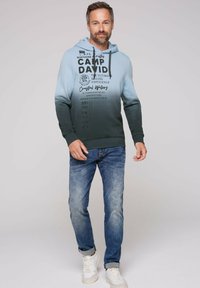 Sudadera con capucha en un degradado de azul claro a verde oscuro con estampado gráfico. Combinada con vaqueros azules y zapatillas blancas.
