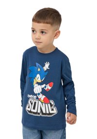 Granatowa koszula z długim rękawem z grafiką Sonica z jeża w czerwono-białych trampkach, a poniżej nadrukowany tekst "SONIC". Wykonana z miękkiej bawełny.
