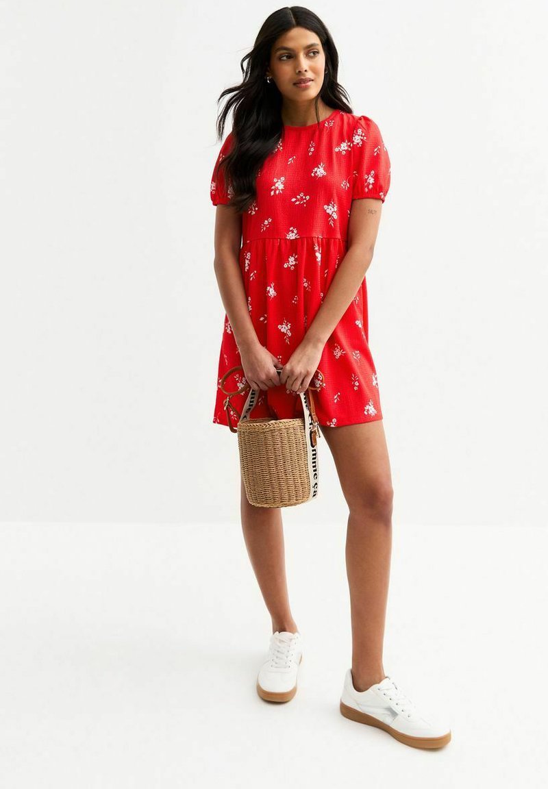 Rotes kurzärmeliges Kleid mit weißem Blumenmuster, ausgestelltes Design, strukturierter Stoff. Das Model hält eine geflochtene Handtasche und trägt weiße Sneakers.