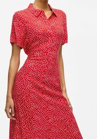 Robe chemise rouge à manches courtes, boutonnée sur le devant, avec des pois blancs. Tissu lisse avec une taille cintrée et une jupe fluide.