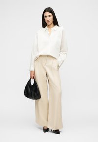 Femme portant une chemise blanche à boutons et un pantalon large beige, tenant un sac à main noir et portant des talons pointus noirs.