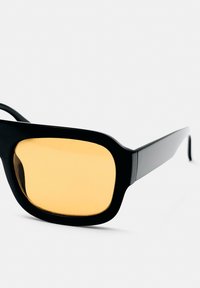 Lunettes de soleil noires avec des montures épaisses et des verres rectangulaires teintés jaunes, vues en gros plan sur un fond blanc.