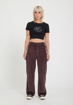 Femme blonde tatouée porte un t-shirt graphique noir court, un pantalon ample en velours côtelé marron et des baskets noires et blanches, debout sur un fond blanc.