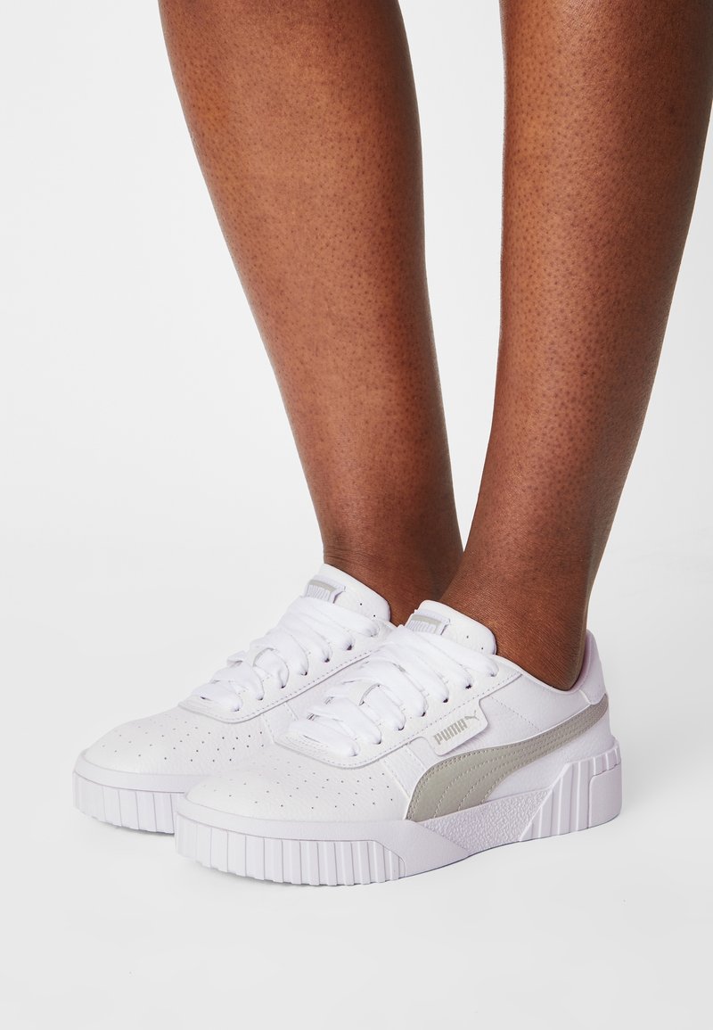 Bílé tenisky s perforovanou texturou, šedými detaily a masivní bílou podrážkou. Obsahují šněrování a logo značky.