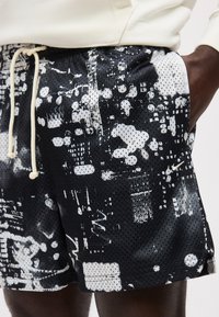 Άτομο που φοράει μαύρα και λευκά σορτς με αφηρημένο μοτίβο από δίχτυ, με μπεζ κορδόνι και το χέρι να ακουμπά στην τσέπη.