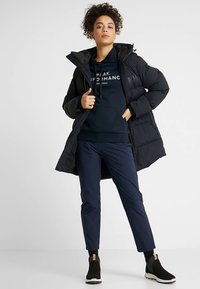 Długa czarna kurtka puchowa z kapturem, noszona na granatowej bluzie z kapturem i granatowych spodniach. Posiada elastyczne mankiety i sięga do kostek, w połączeniu z czarnymi butami wsuwanymi.