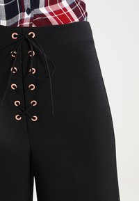 Svarta högmidjade byxor med en snörningsdesign framåtriktad med kopparfästen och matchande snören, tillverkade av mjukt, texturerat tyg.