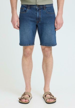 Mann iført midtlange blå denimshorts og beige sandaler med dobbelt reim, stående med avslappede hender langs siden.