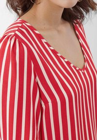 Rote und cremefarbene gestreifte Bluse mit V-Ausschnitt, die angesprachte Ärmel und eine weiche Stofftextur aufweist. Beachten Sie die Kontraststreifen für visuelle Details.