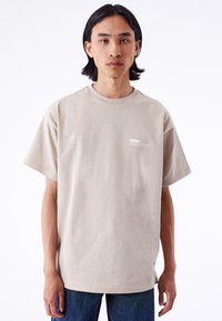 Camiseta de algodón beige con mangas cortas, corte relajado, escote redondo y pequeño logo blanco impreso en el lado izquierdo del pecho.