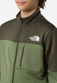 Roheline fliisjope lukuga rinnatasku, kõrge krae ja valge logoga vasaku ülaosas. Omab tekstuuriga õlapaneele.