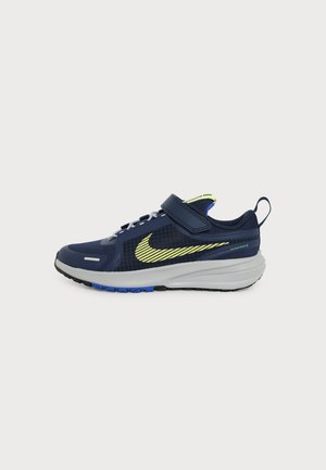 Chaussure de sport bleu foncé avec le swoosh Nike jaune, semelle blanche, semelle extérieure bleue et une bride velcro unique sur les lacets, sur un fond clair uni.