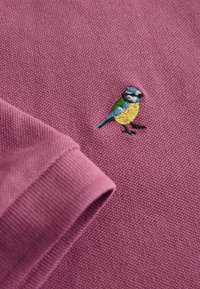 Tissu texturé violet avec une manche pliée et un petit oiseau brodé avec des fils bleu, jaune, vert et noir.