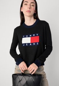 Un mannequin portant un pull noir avec un logo "TOMMY JEANS" se tient debout, tenant un sac à main matelassé noir, sur un fond neutre.