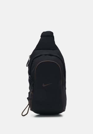 Sort Nike skuldertaske med lynlåslomme foran, buet design og justerbar rem, vist mod en ensfarvet grå baggrund.
